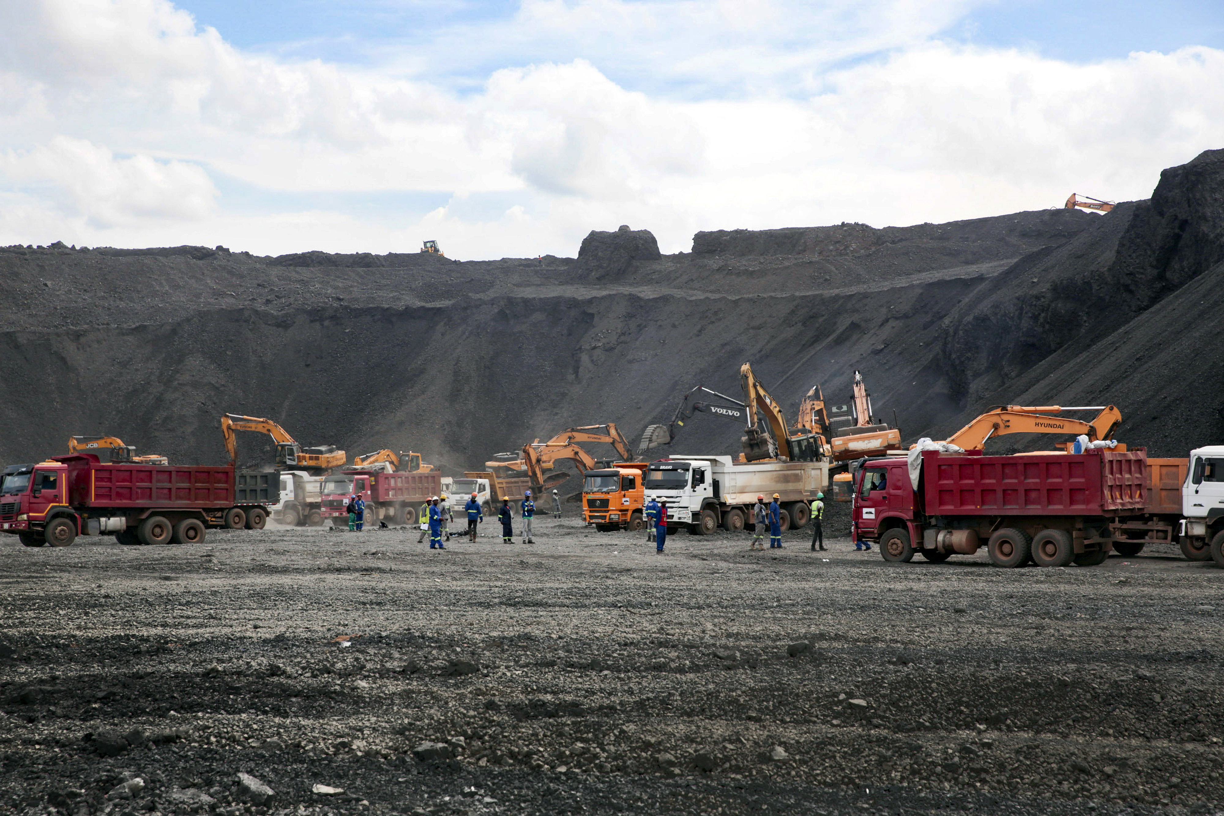 Schlackehalde der Mopani Glencore Kupfermine in Sambia im Januar 2019. Die Schlacke enthält noch Erze und Reste von Kupfer. Viele Familien suchen auf der Halde (illegal) nach Rohstoffen, um damit ein Einkommen zu erzielen. Die Halde ist im Hintergrund zu sehen. Im Vordergrund stehen Lastwagen und Bagger, dazwischen stehen Arbeiter.