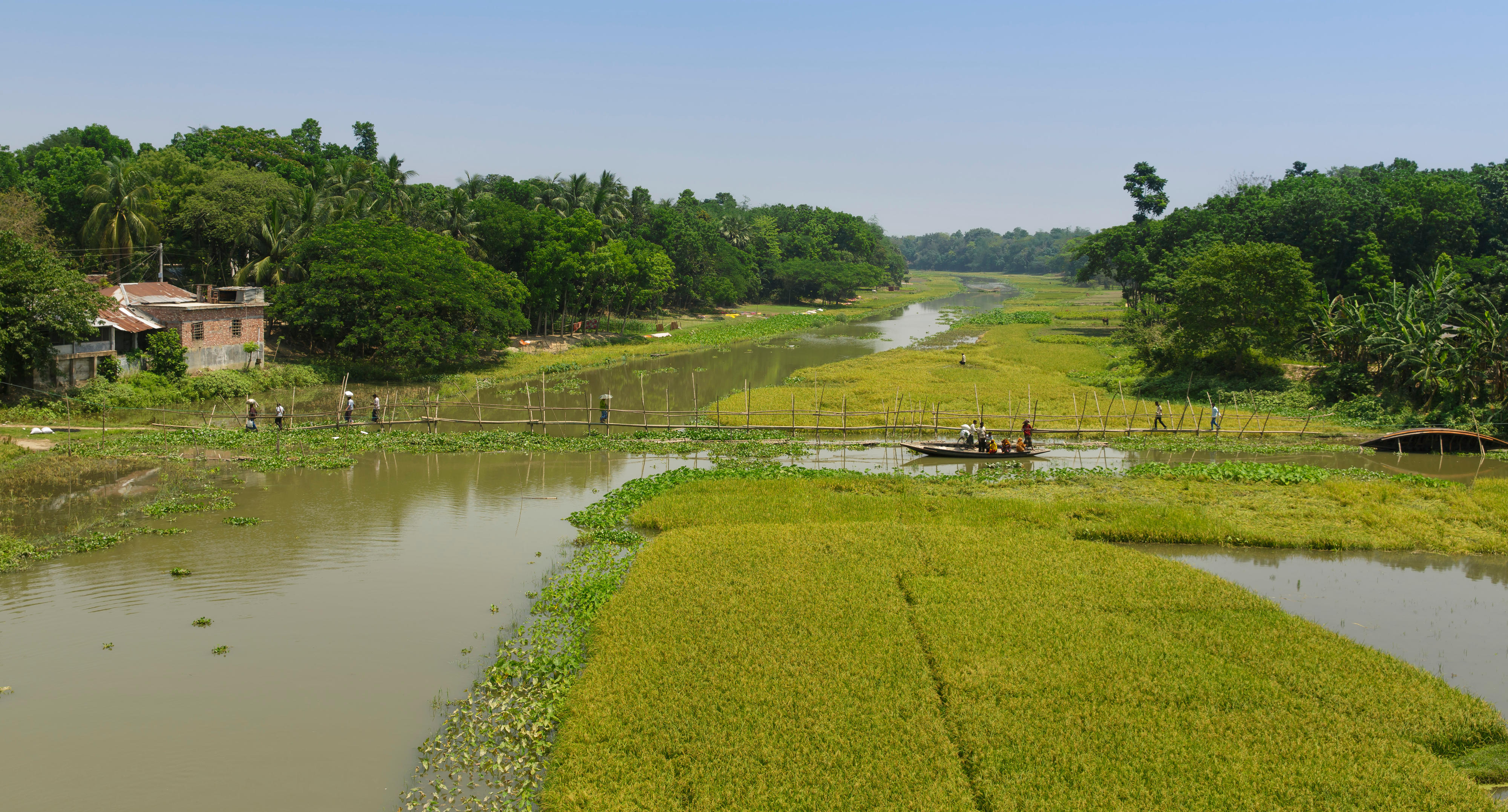 Menschen überqueren über eine schmale Fußgängerbrücke und per Boot einen Fluss in Bangladesch.