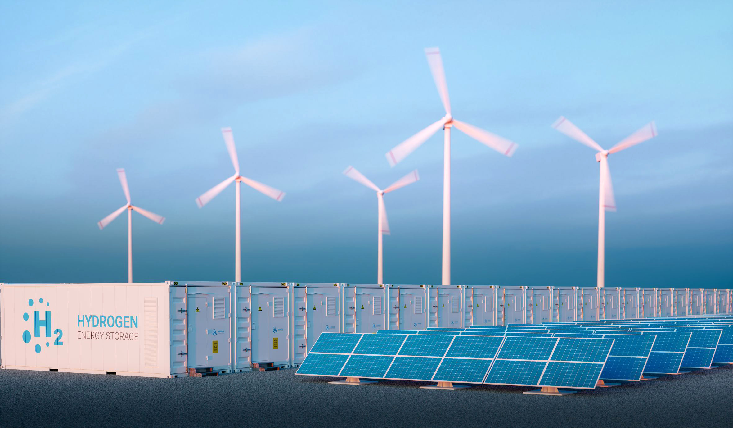 Symbolbild: Gewinnung von grünem Wasserstoff. Im Vordergrund Solarzellen, dahinter Container zur Speicherung von Wasserstoff, im Hintergrund Windkraftanlagen mit drehenden Rotoren