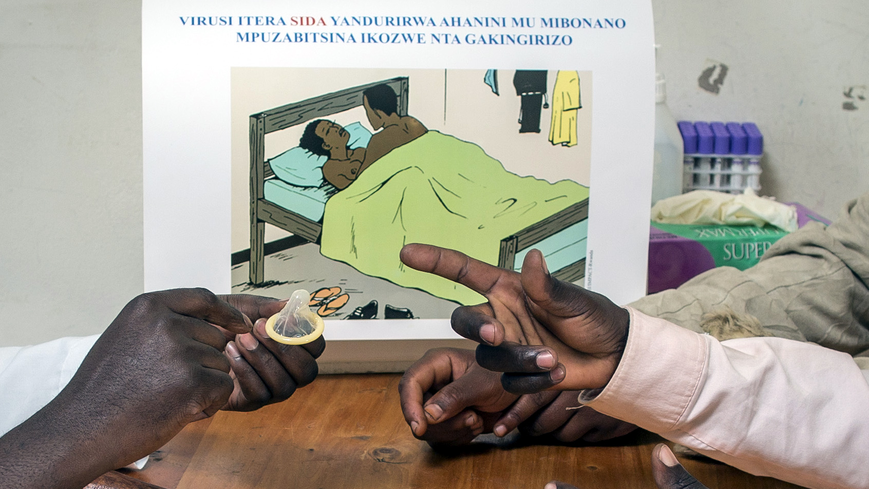 Instructions on condom use Detail photograph: arms of people sitting around a desk, with two hands on the left holding a condom and two arms and hands visible on the right