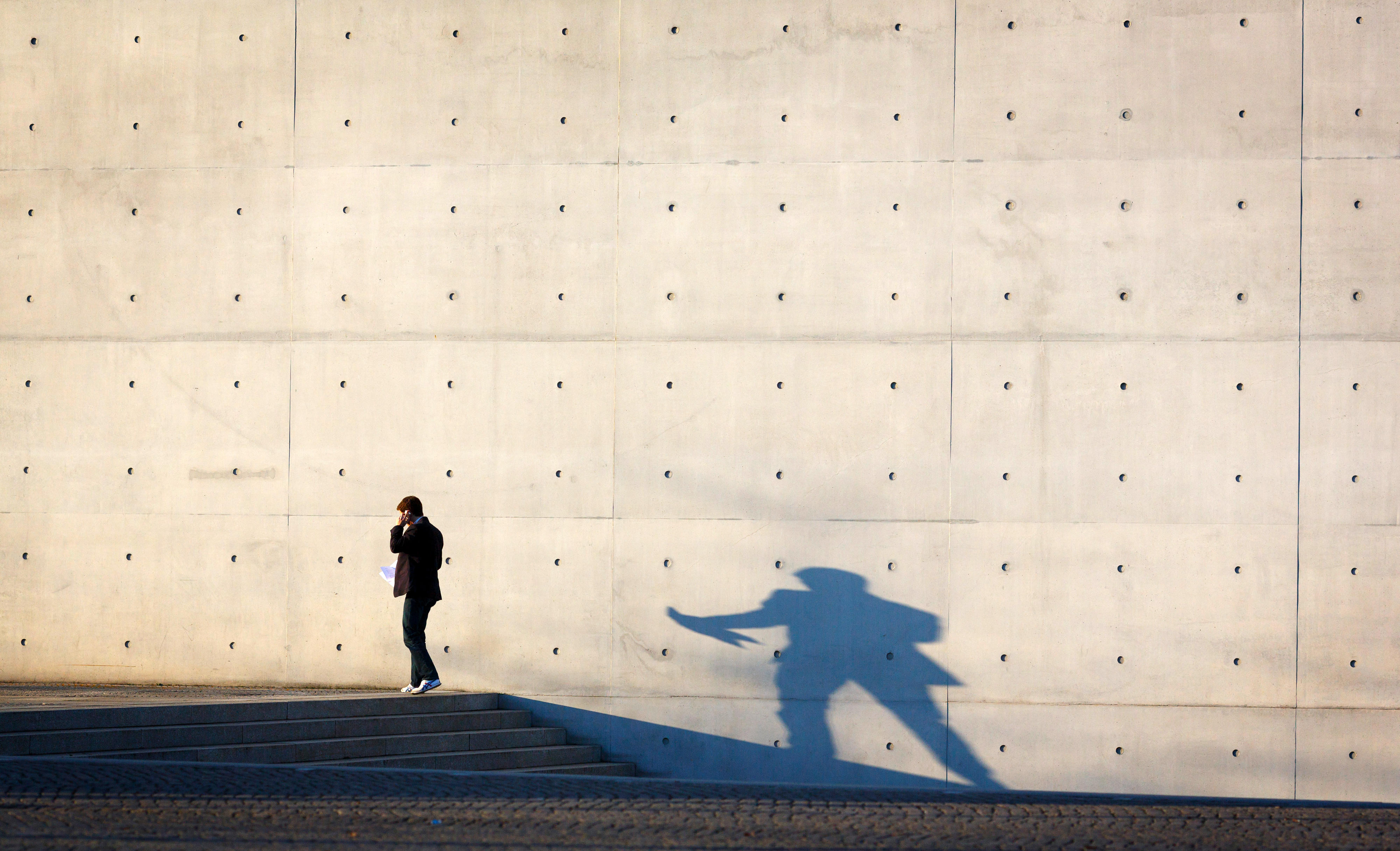 A person is talking on the phone in front of a concrete wall, casting a shadow.
