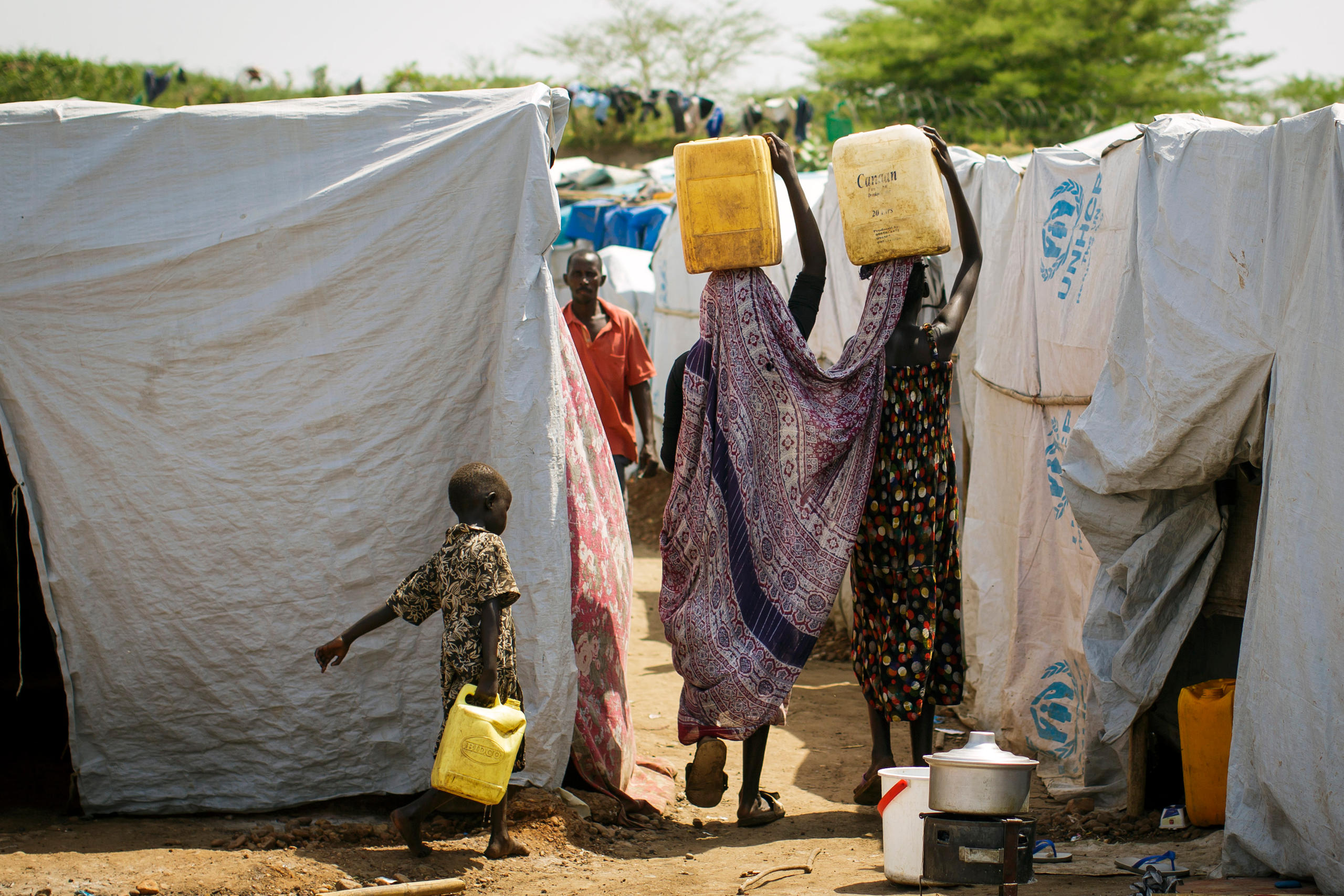 Foto von Frauen, die Kanister auf ihrem Kopf tragend zwischen Zelten laufen.