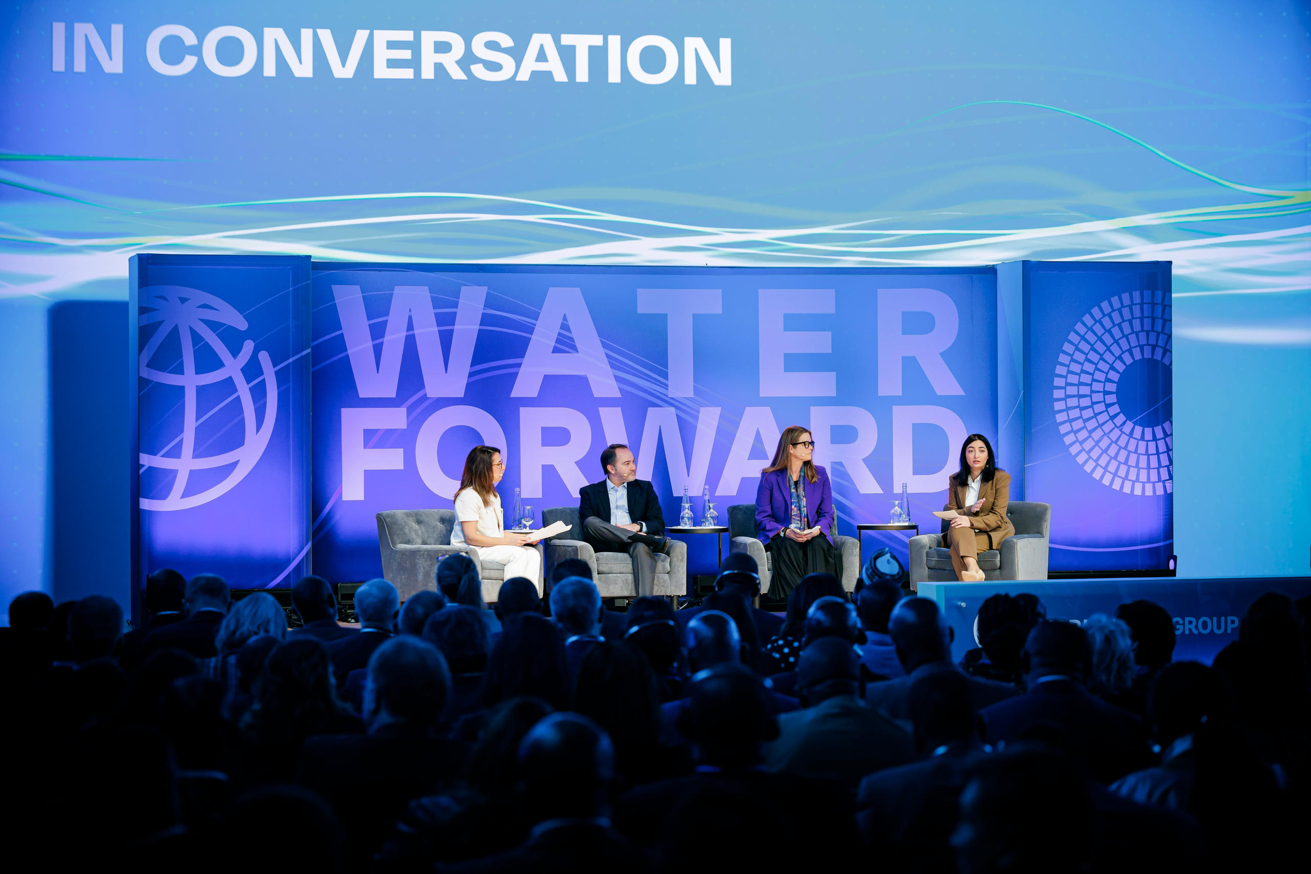 Foto von vier Menschen, die auf einer Bühne sitzen. Im Hintergrund ein Plakat, auf dem Water Forward steht.