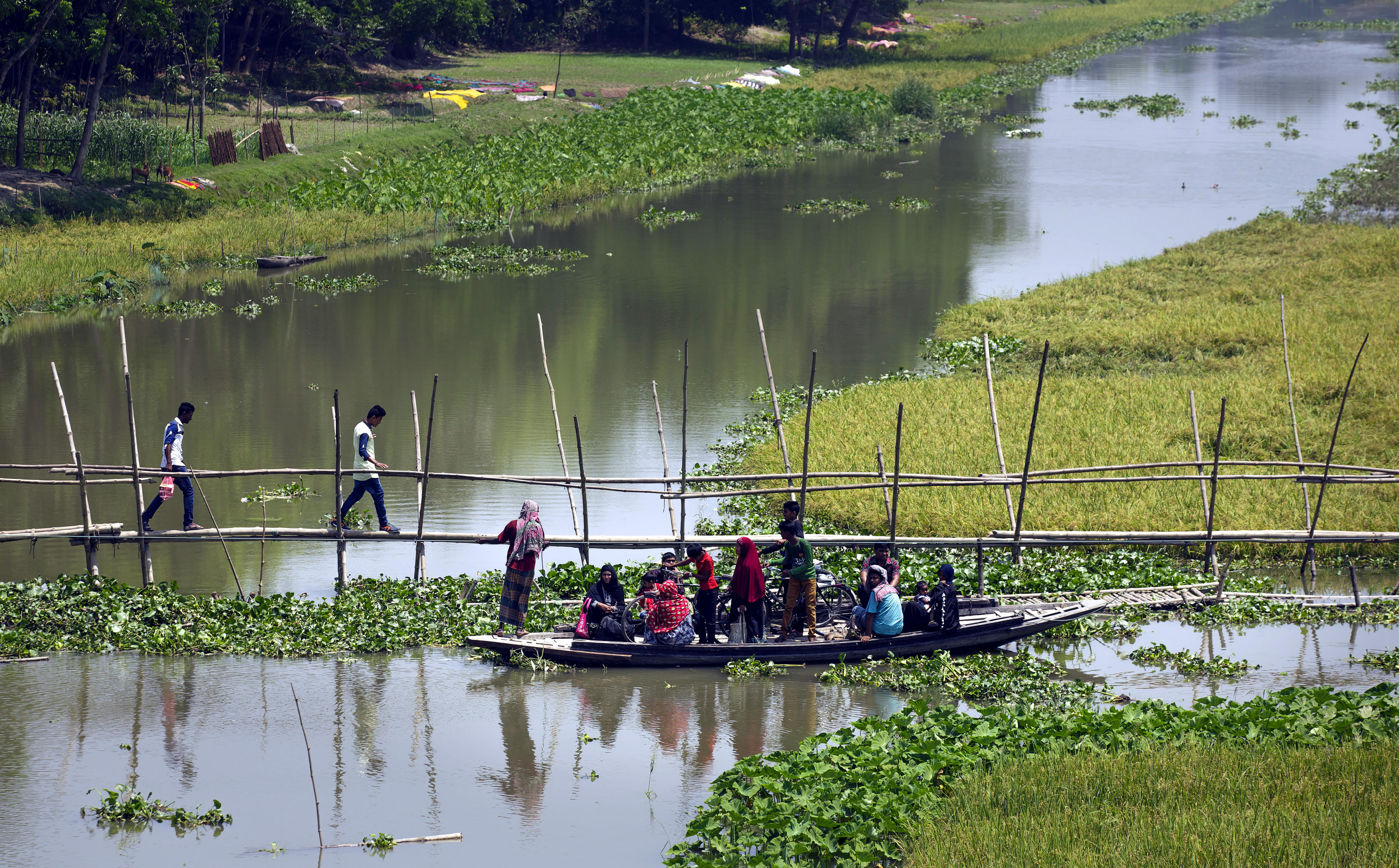 Menschen überqueren über eine schmale Fußgängerbrücke und per Boot einen Fluss in Bangladesch.