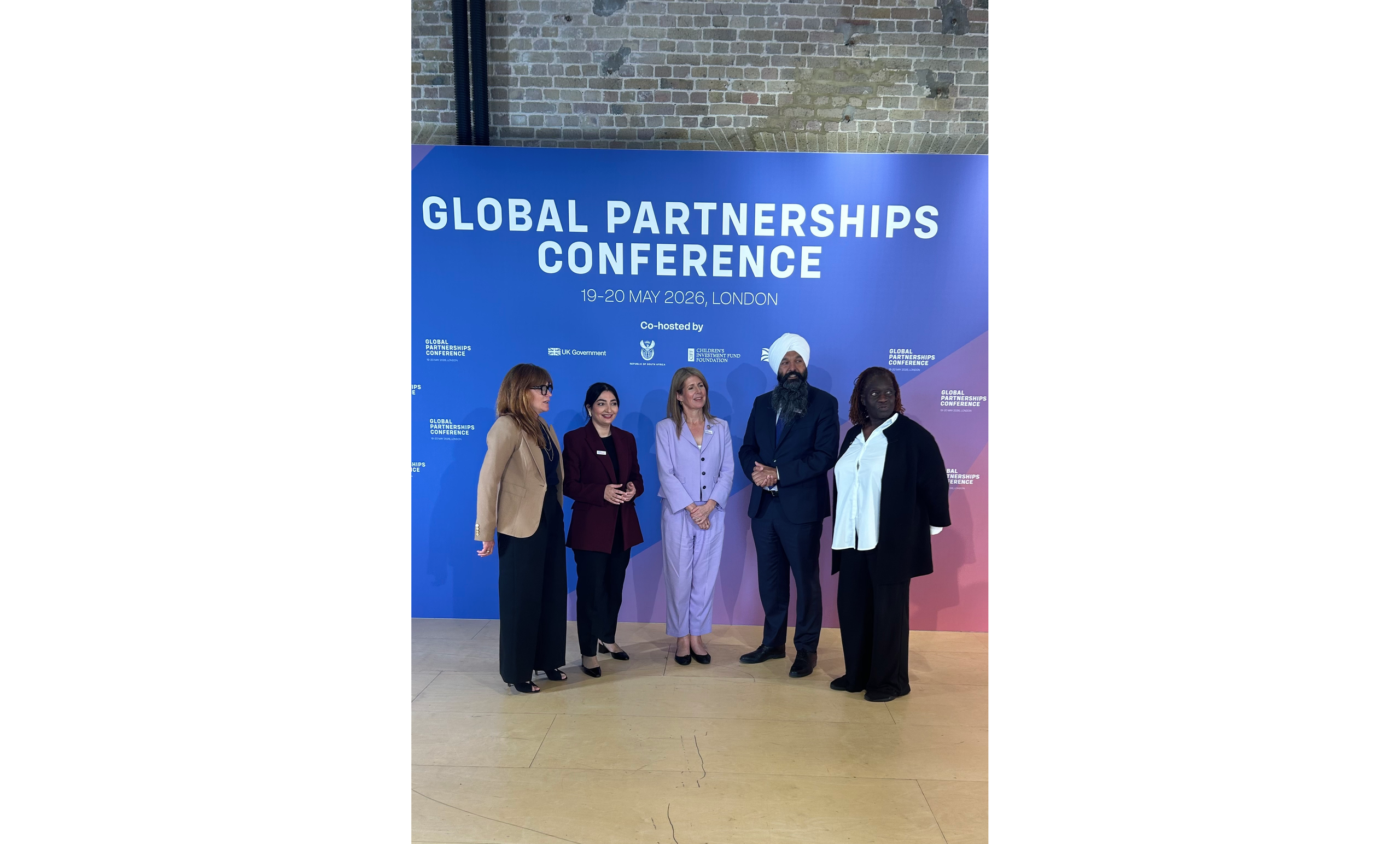 Foto von fünf Menschen, die nebeneinander stehen. Hinter ihnen ein Plakat, auf dem "Global Partnership Conference" steht.