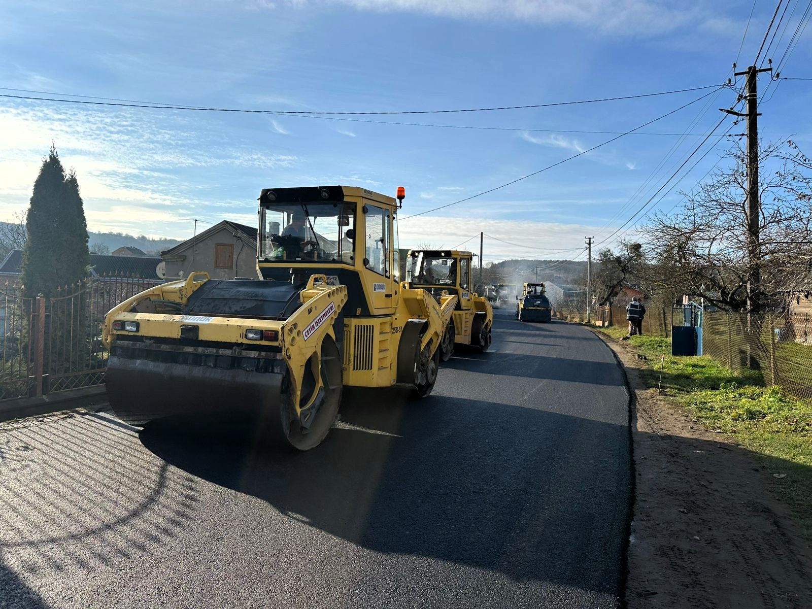 Straßenwalzen auf frisch asphaltiertem Landweg mit Häusern und Bäumen im Hintergrund.
