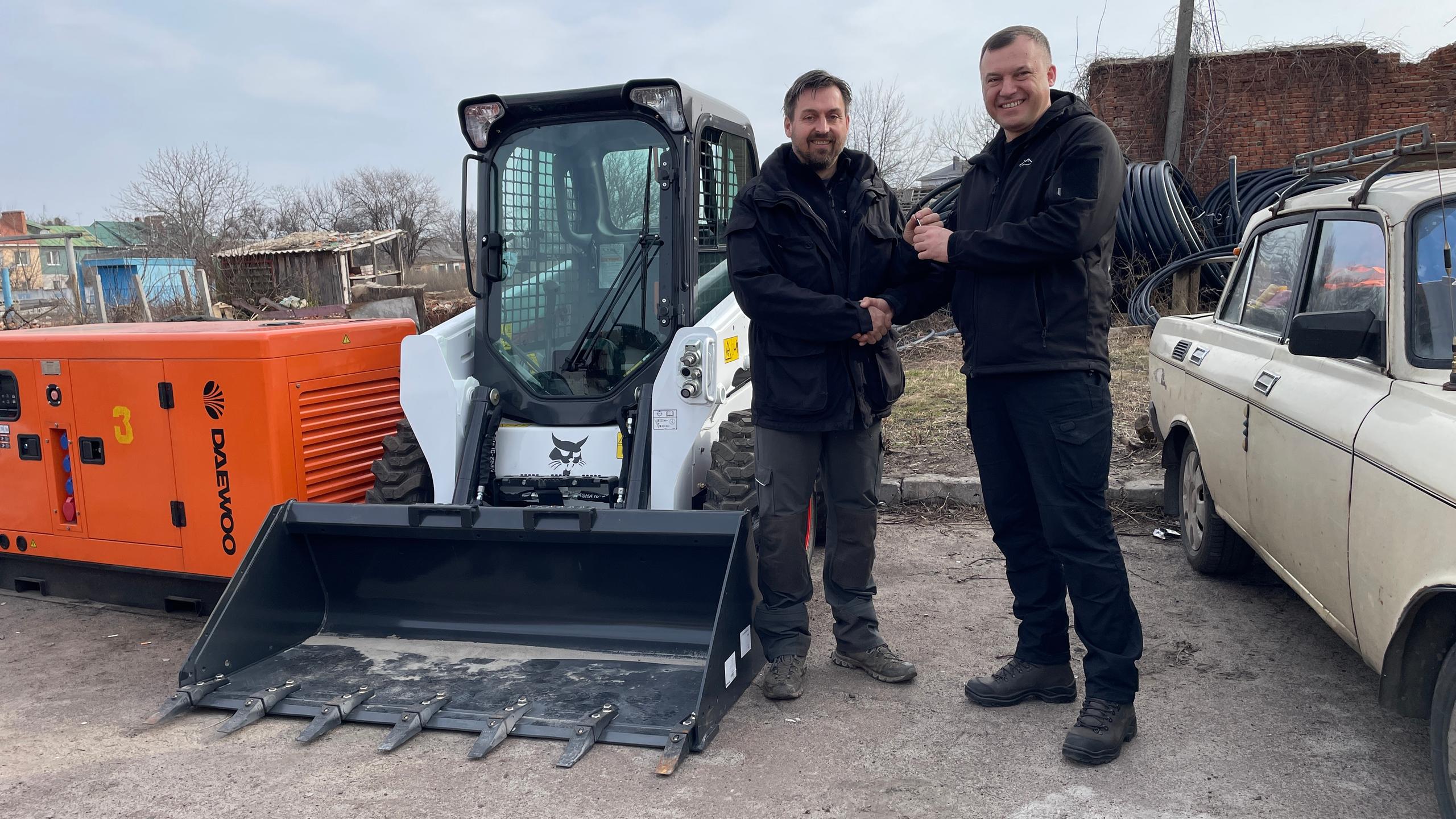 Zwei Männer in schwarzer Arbeitskleidung stehen vor einem weißen Bobcat-Kompaktlader und schütteln sich die Hand.