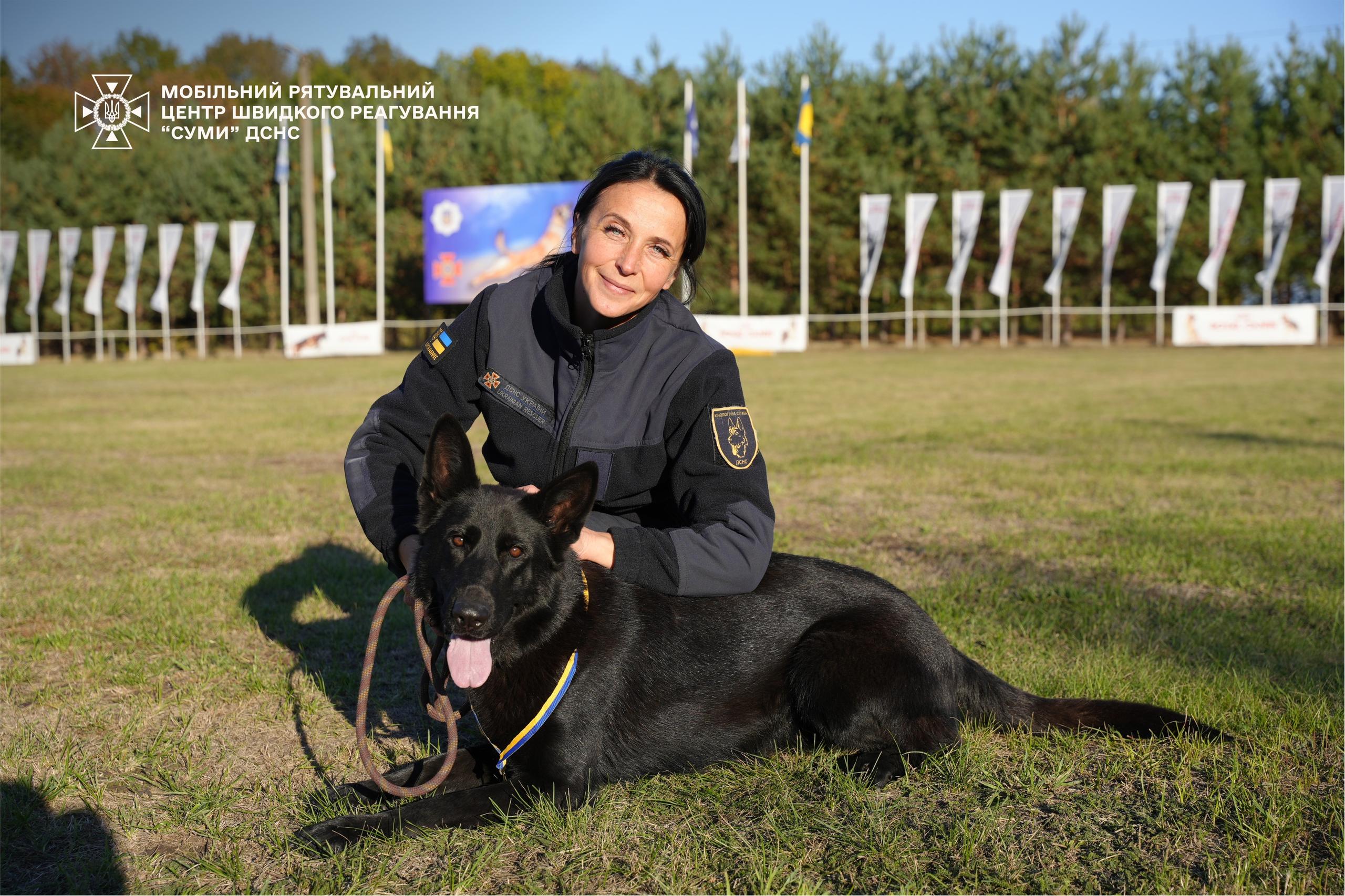 Eine Frau in schwarzer Arbeitskleidung streichelt einen schwarzen Schäferhund.