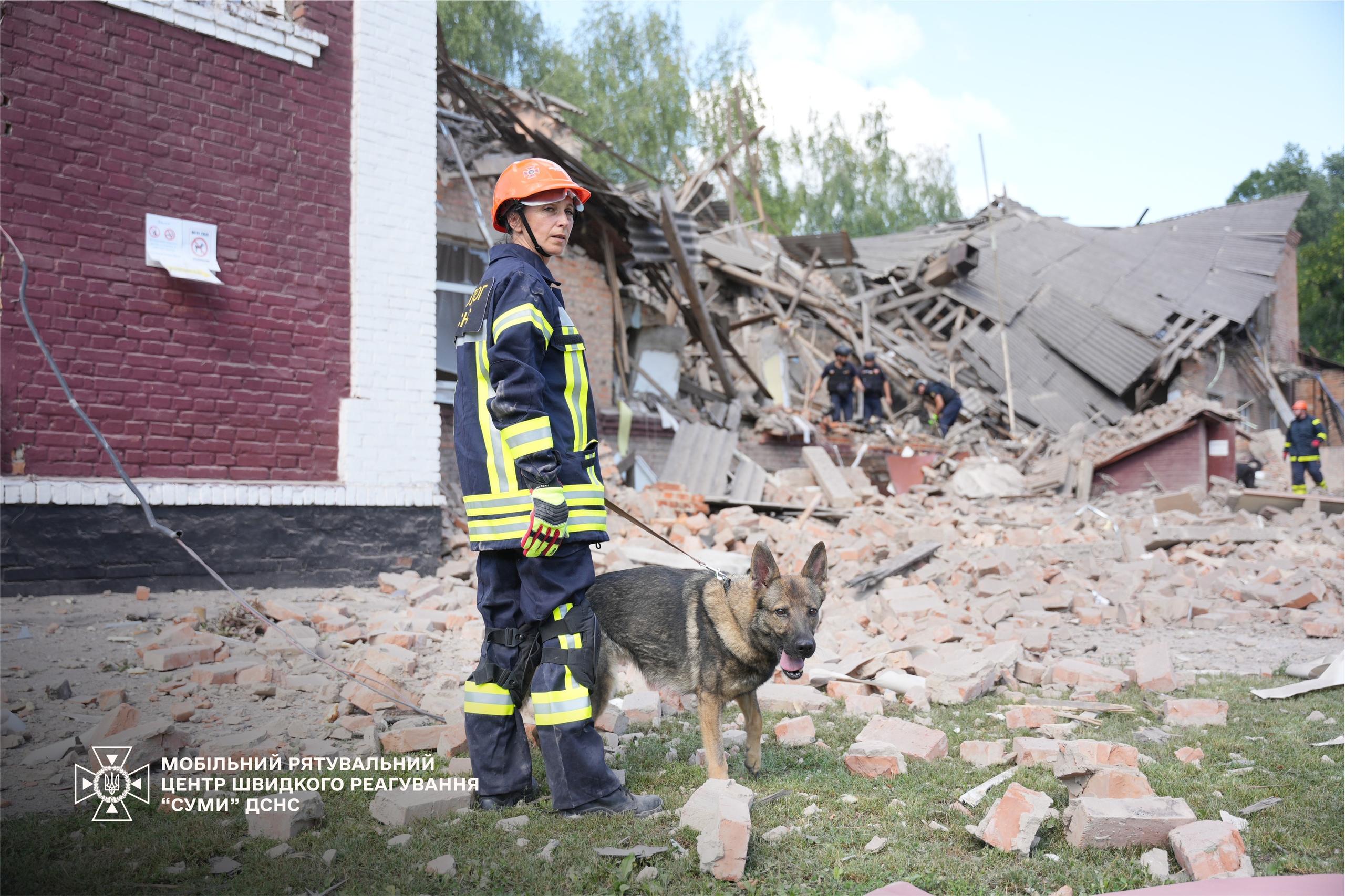 Frau in Schutzkleidung und Helm neben einem Schäferhund vor Trümmern.
