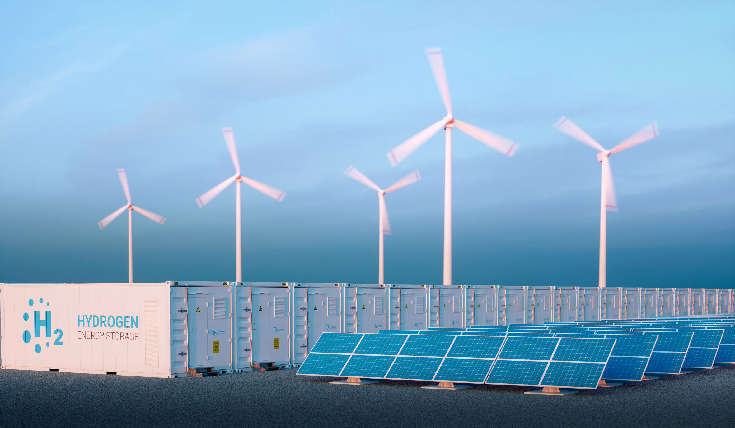 Illustrative image: Production of green hydrogen. In the foreground are solar panels; behind them are containers for storing hydrogen; in the background are wind turbines with rotating rotors.