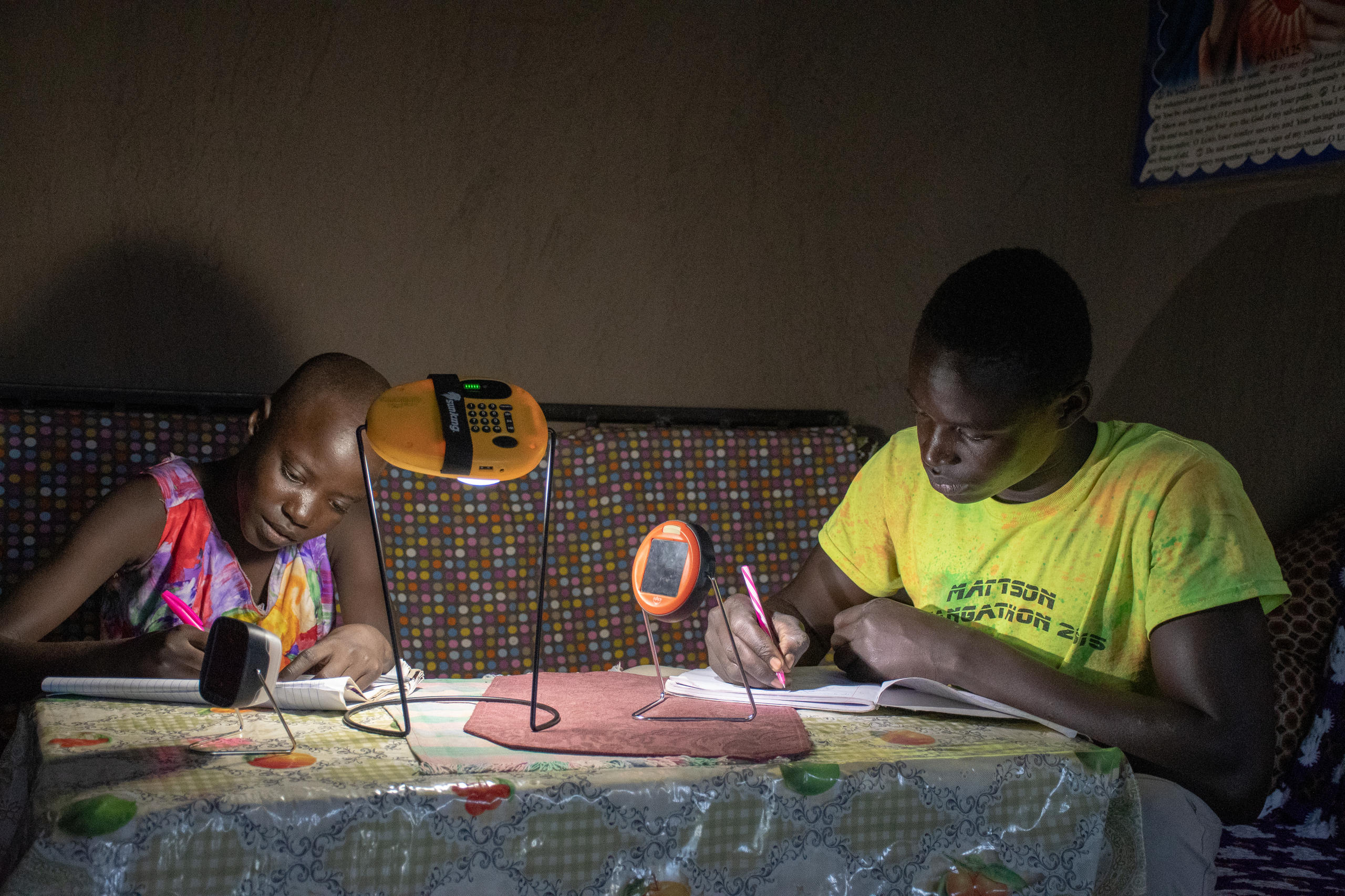 A photo of two people sitting at a table and writing by the light of a lamp.