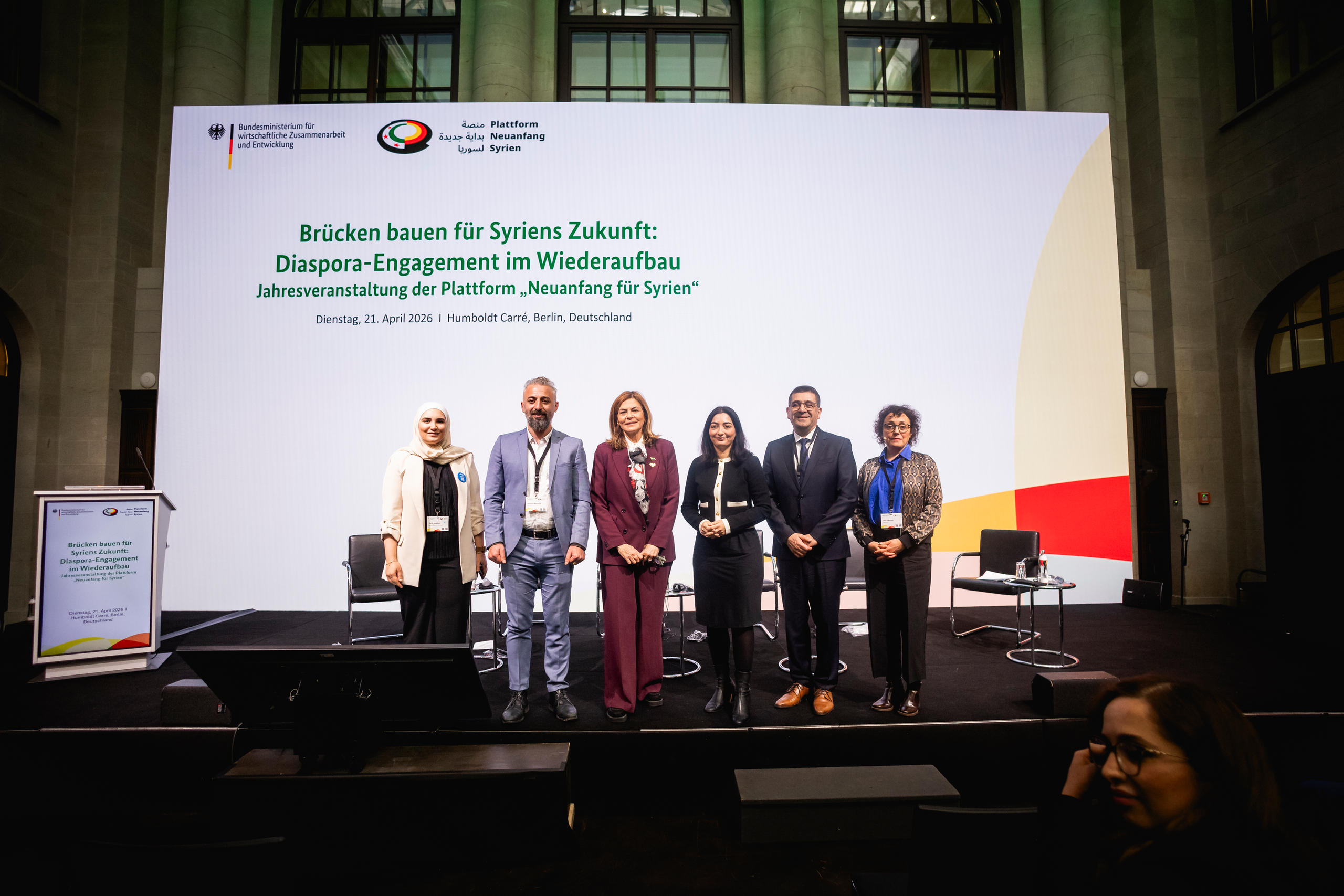 Five people are standing side by side on a stage in front of a large screen displaying text about the diaspora engagement event for Syria.