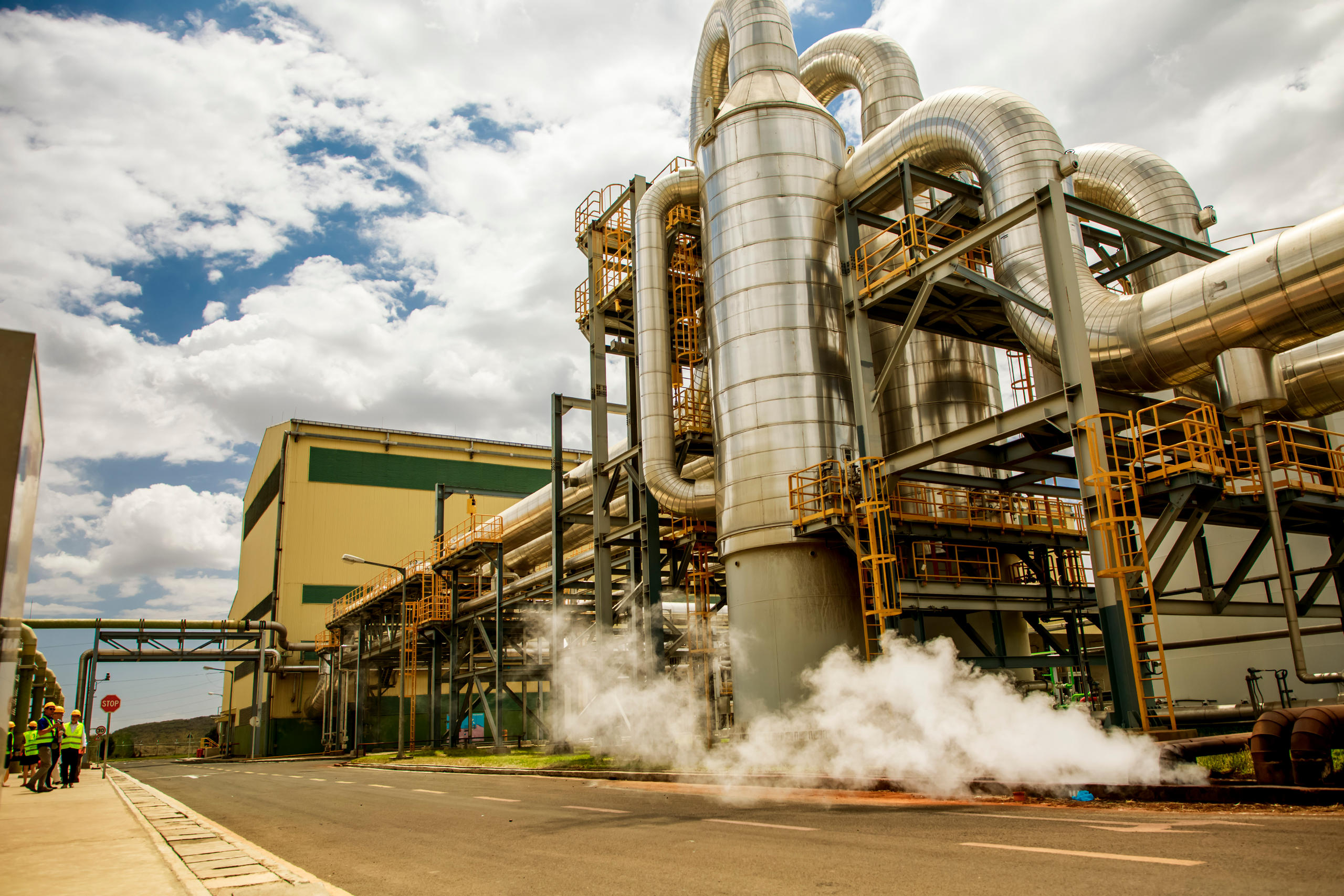 Industrielle Anlage mit großen silbernen Rohrleitungen und Dampfaustritt vor gelb-grünem Gebäude unter bewölktem HimmelIndustrielle Anlage mit großen silbernen Rohrleitungen und Dampfaustritt vor gelb-grünem Gebäude unter bewölktem Himmel.