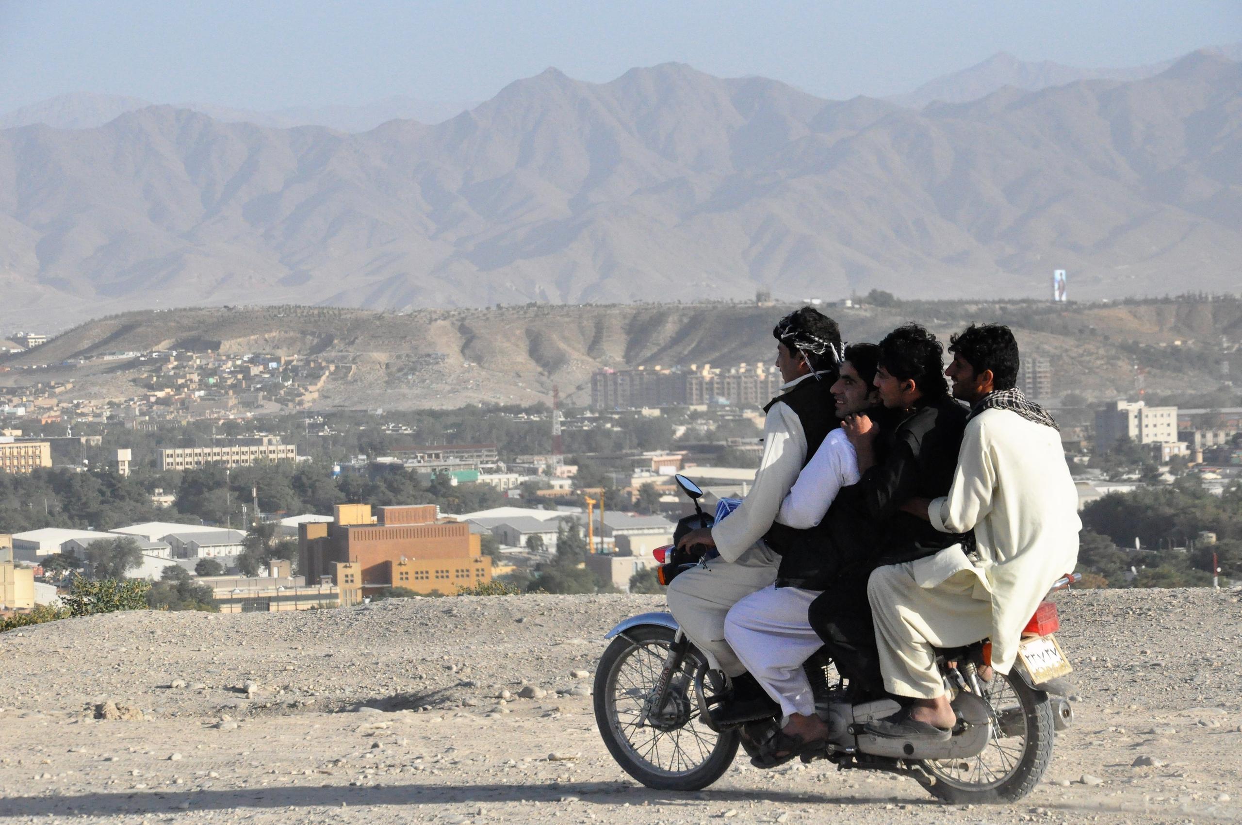 Four men wearing traditional clothing riding a motorcycle on a dirt hill overlooking a city with mountains in the background