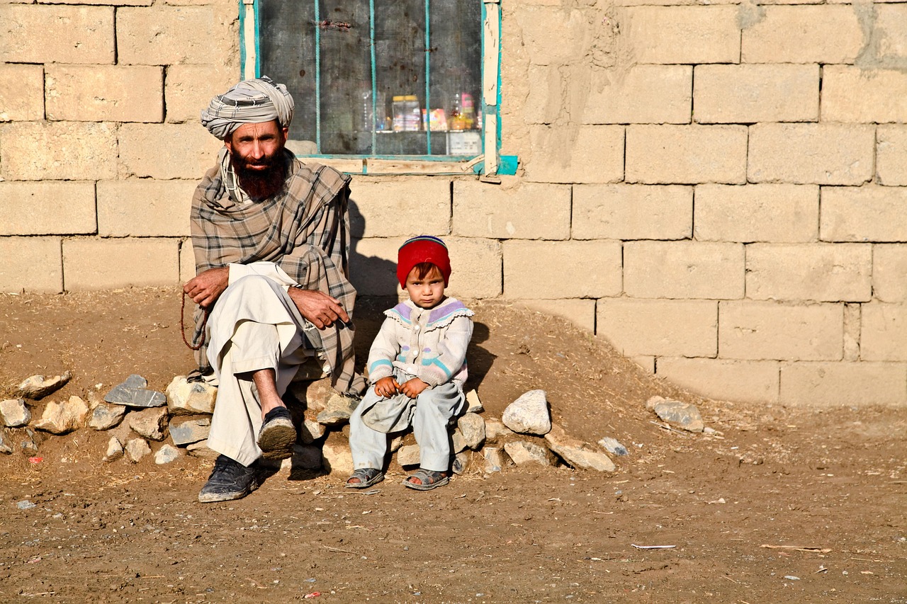 Man wearing traditional clothing and turban sitting next to a young child in a red hat outside a stone building.