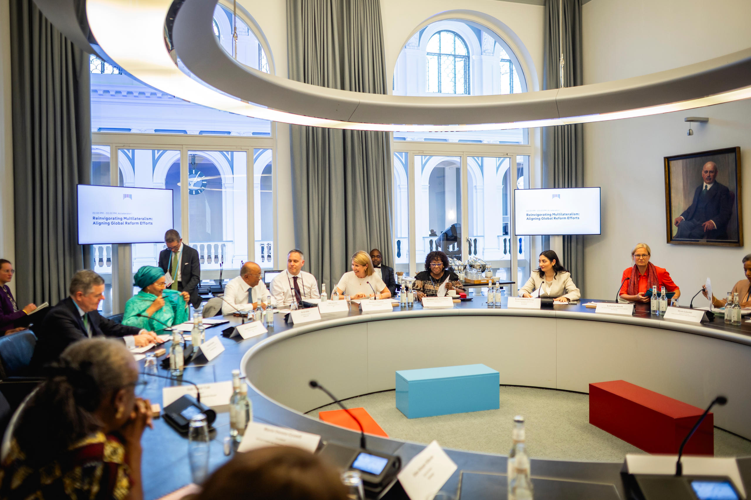 Menschen sitzen an einem runden Konferenztisch in einem Raum mit großen Fenstern und zwei Bildschirmen an der Wand.