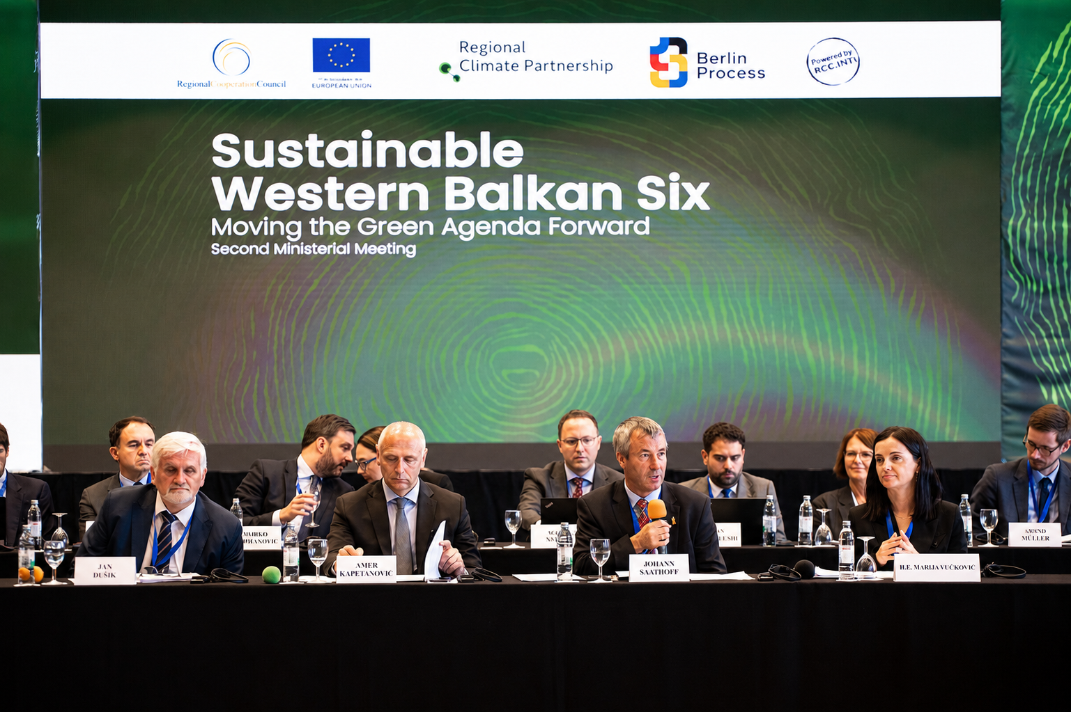 Mehrere Menschen sitzen an Tischen vor einer Wand auf der Sustainable Western Balkan Six steht. Ein Mann hat ein Mikrophon in der Hand.