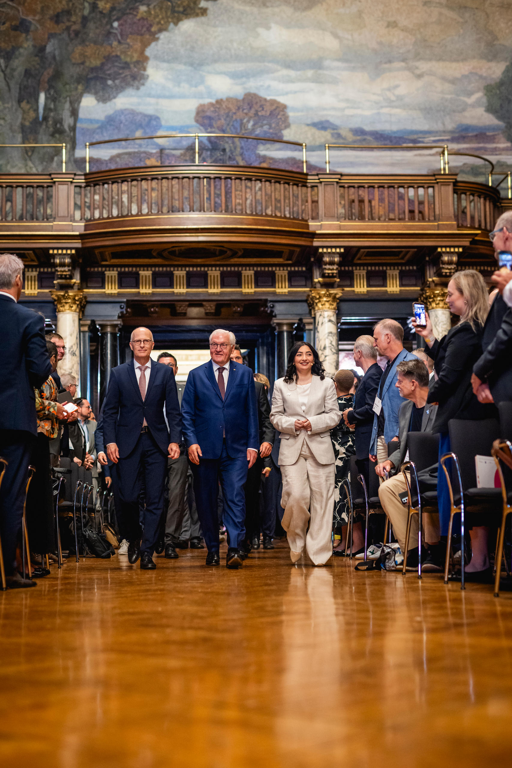Drei Menschen laufen auf die Kamera zu, rechts und links sitzen und stehen Zuschauer.