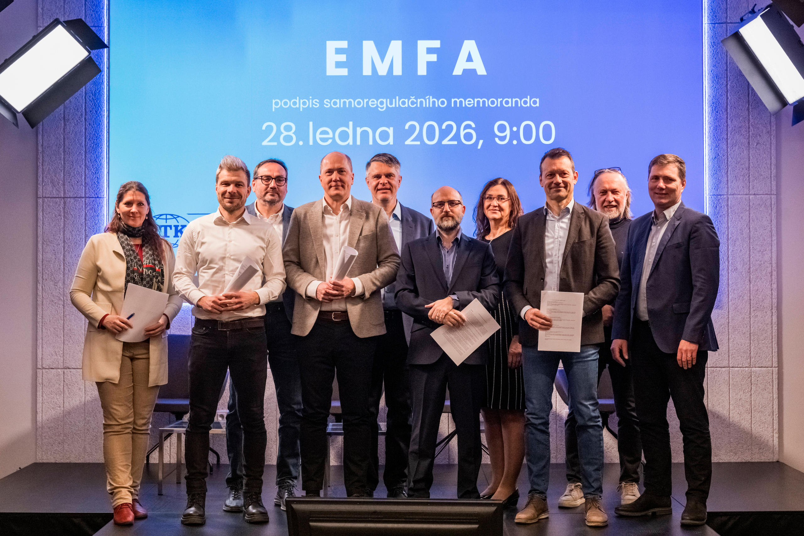 Gruppe von neun Personen steht vor einer Wand mit der Projektion 'EMFA podpis samoregulacního memoranda 28. ledna 2026, 9:00'.