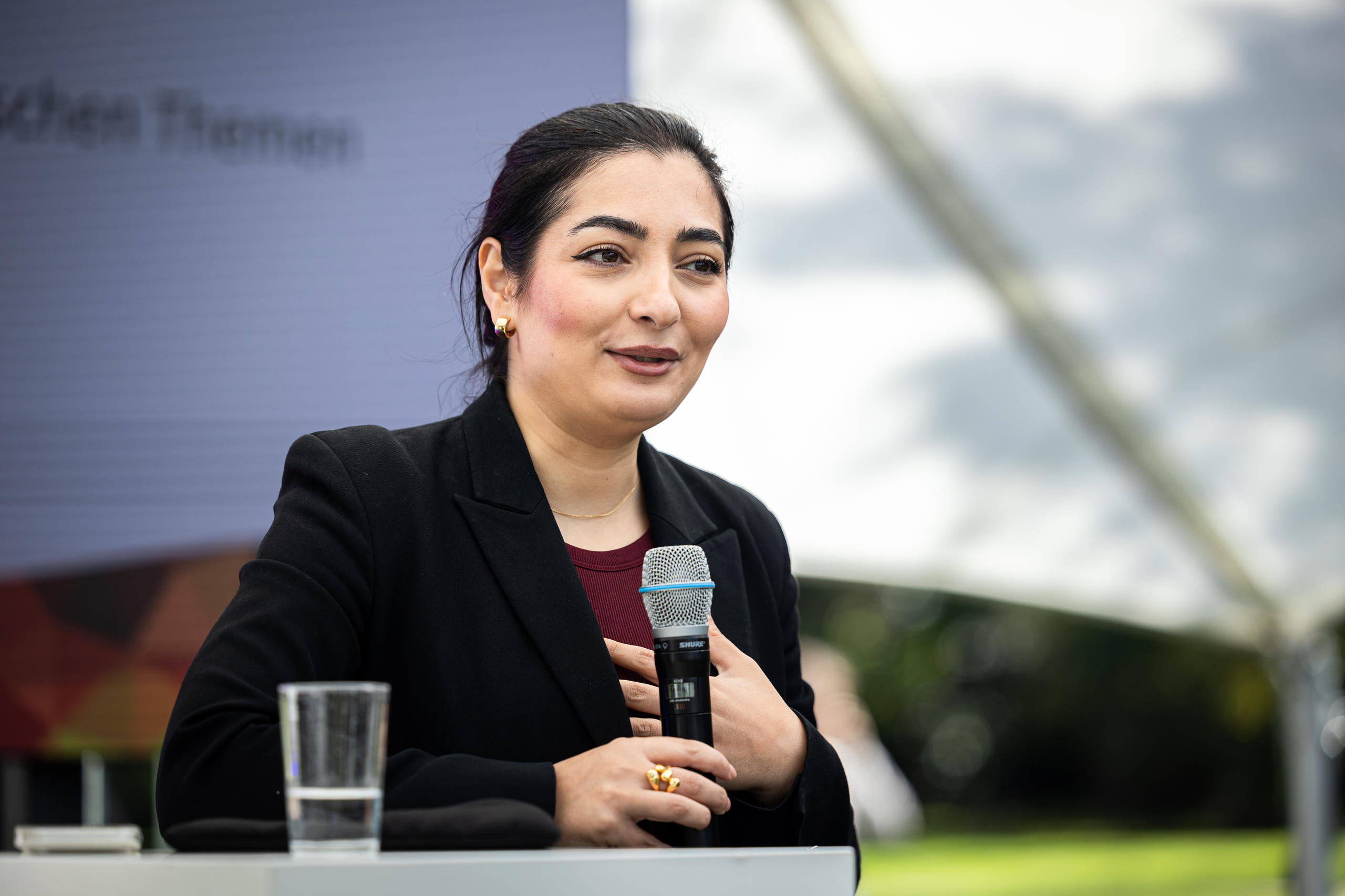 schreibe einen alttext für dieses bild Eine Frau in schwarzem Blazer spricht bei einer Veranstaltung in ein Handmikrofon.