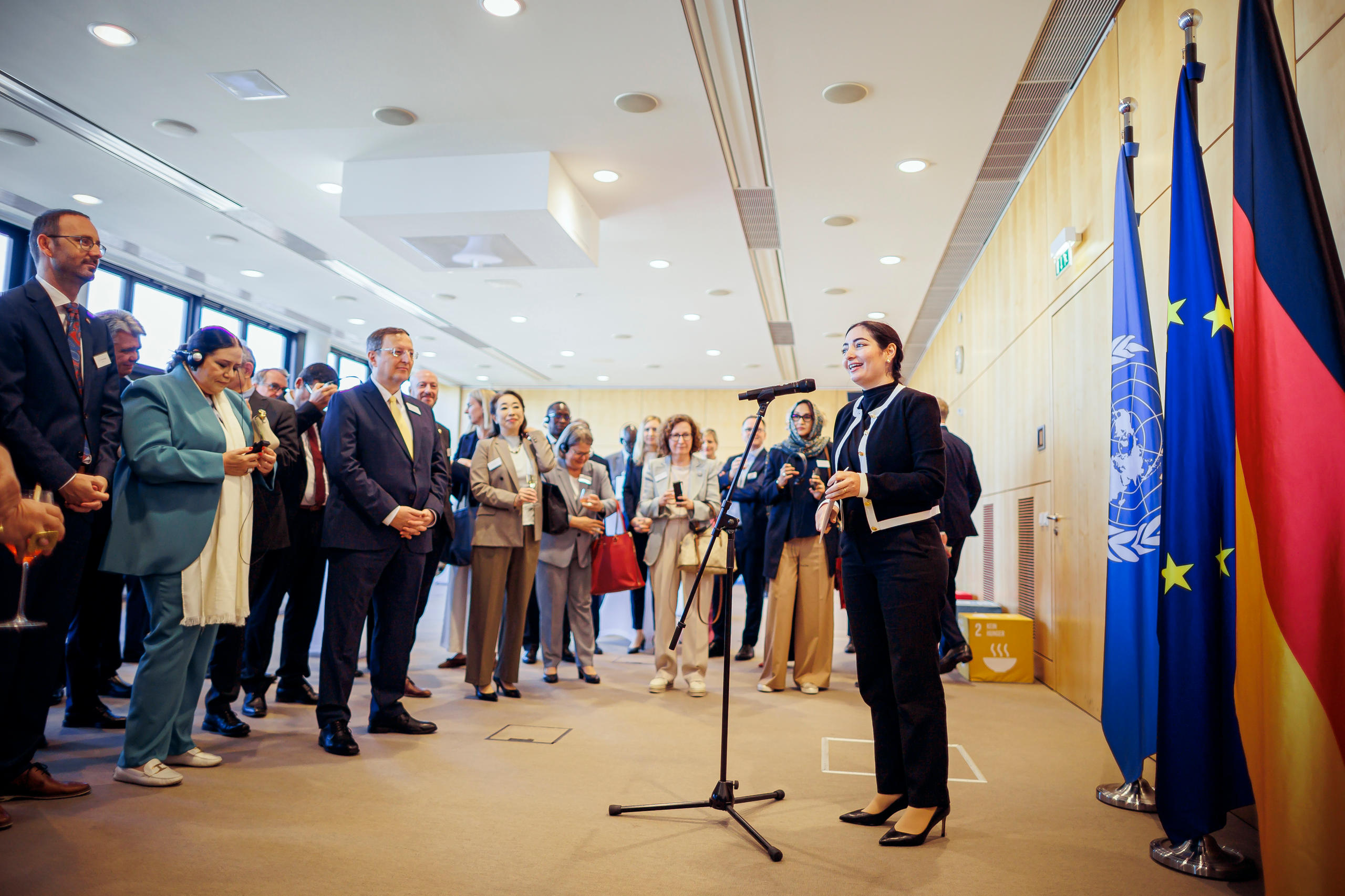 Eine Frau spricht vor einer Gruppe von Gästen an einem Mikrofon. Neben ihr stehen die Flaggen der Vereinten Nationen, der Europäischen Union und Deutschlands.