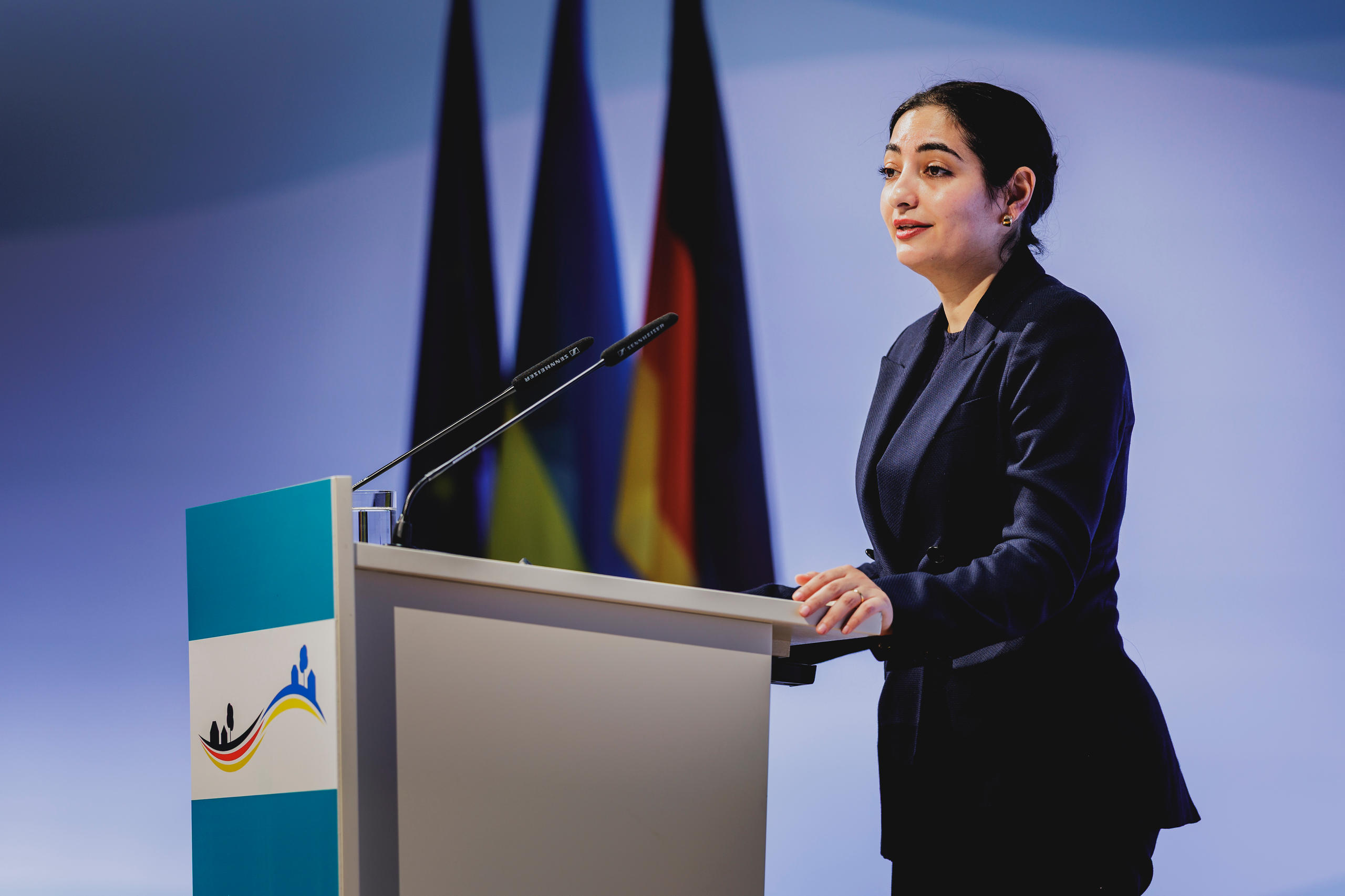 Eine Frau hält an einem Rednerpult eine Rede; hinter ihr stehen mehrere Flaggen, darunter die deutsche Flagge.