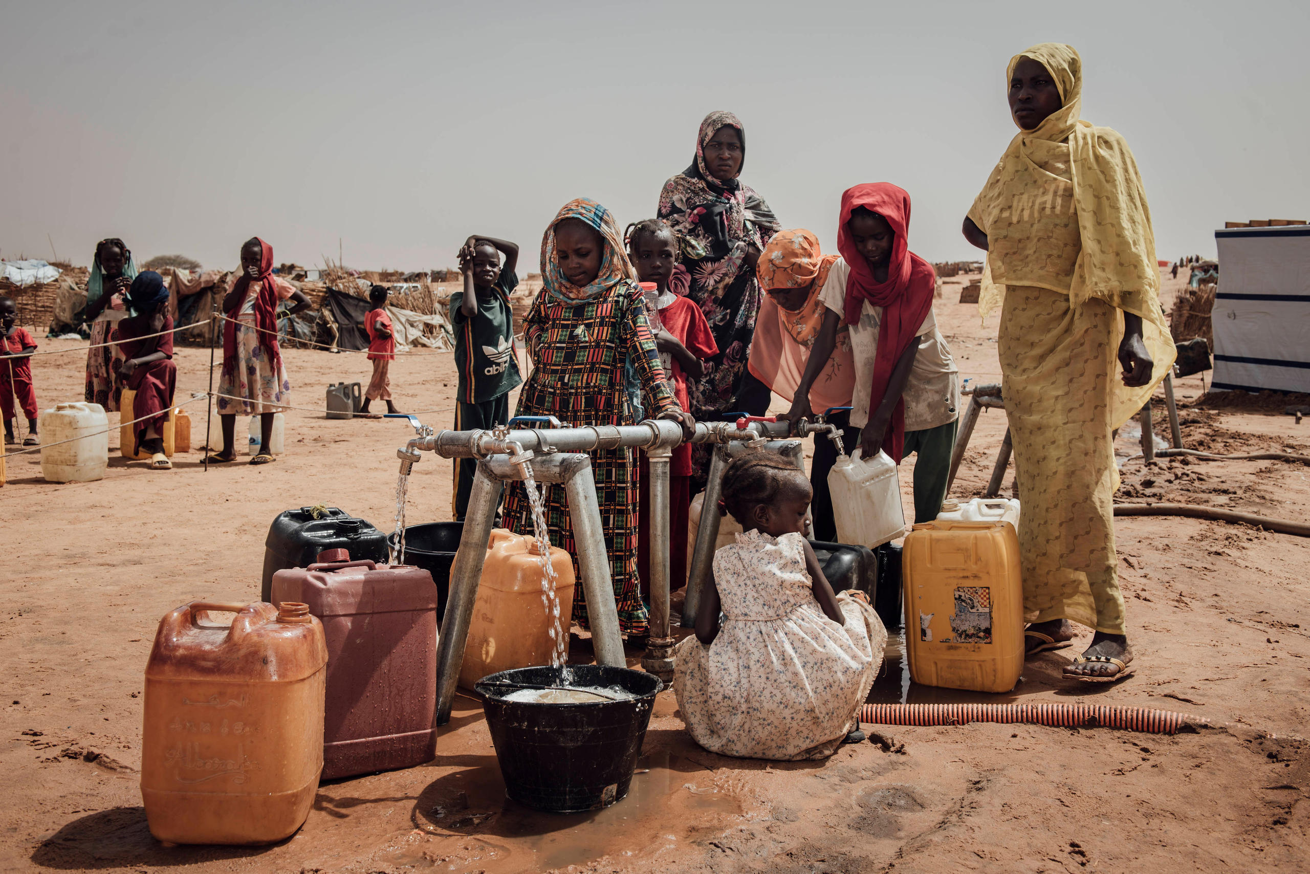 Mehrere Frauen und Kinder stehen an einer Wasserstelle mit Kanistern und einem Wasserhahn in einer trockenen, sandigen Umgebung.