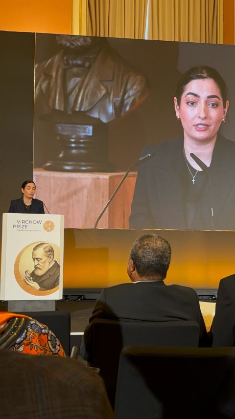 Woman speaking at a podium with a bust sculpture behind her and her image projected on a large screen in a conference setting.