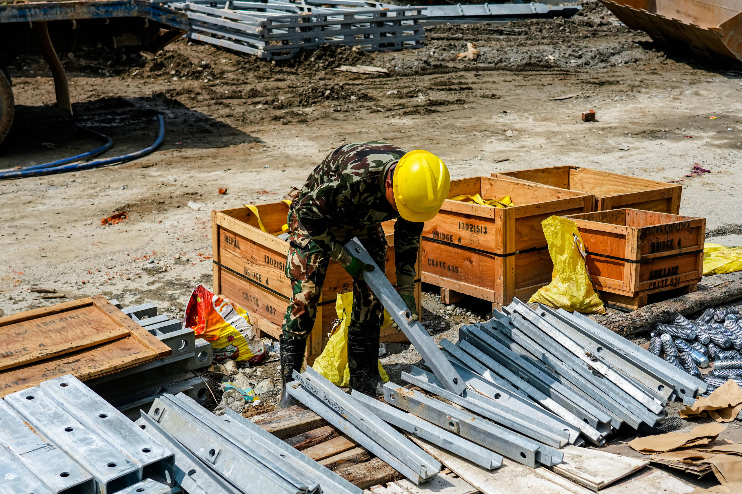 Ein Mann hebt auf einer Baustelle ein Stahlteil zwischen Holzkisten und gestapelten Brückenteilen auf.