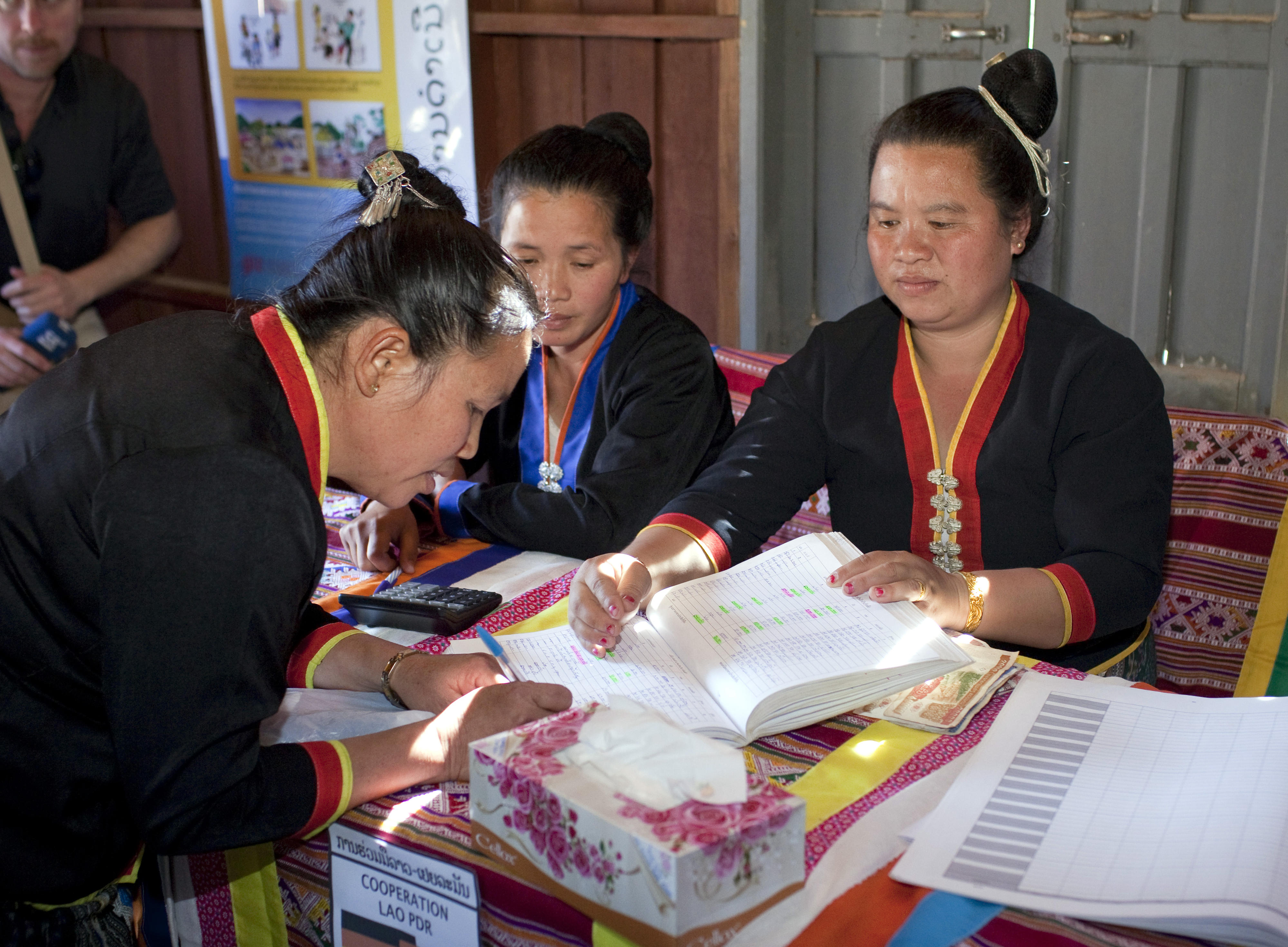 Laos: Am monatlichen Banktag zahlen Mikrofinanzkundinnen Geld beim Dorfbankenkomitee der Dorfbank in Pung ein. Zwei Frauen in traditioneller Kleidung sitzen hinter einem Tisch, eine weiterer steht davor und unterschreibt in einem Buch.