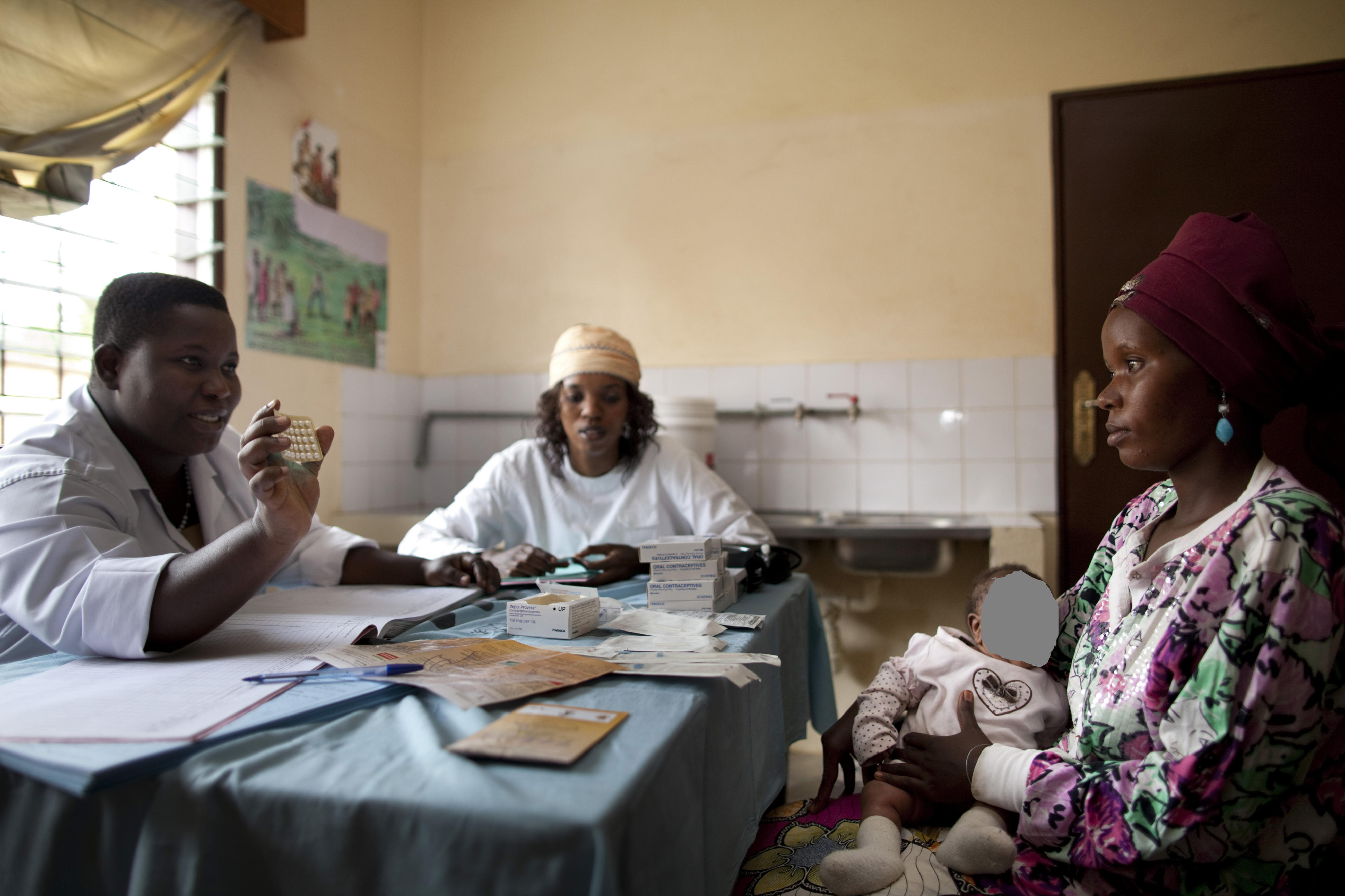 Beratung zum Thema Verhütungsmittel in einer Gesundheitsstation in Bujumbura, Burundi. Eine Frau mit einem Kind auf dem Schoß sitzt am Tisch. Ihr gegenüber sitzt eine Beraterin in einem weißen Kittel und zeigt ihr eine Tablettenpackung.