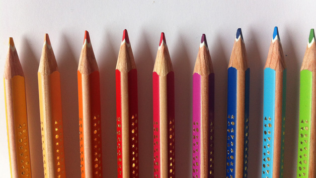 Symbolbild: Verschiedenfarbige Buntstifte liegen in einer geraden Reihe nebeneinander auf einem weißen Hintergrund